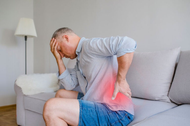 middle aged man holding head and lower back in pain on couch